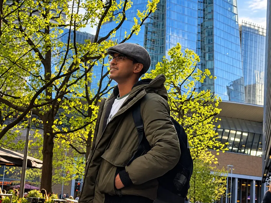 Faraz Ansari in a green jacket and flat cap, looking up, with the Shard and glass office towers behind him